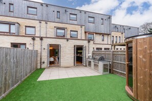 A modern two-story building with a patio area featuring artificial grass, a barbecue station, and a wooden fence.