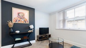 A modern living room with a dark accent wall, a flat-screen TV, a glass coffee table, and a window with blinds. - Modern Apartment By Historic Gloucester Quays (Gloucestershire)