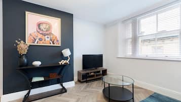 A modern living room with a dark accent wall, a flat-screen TV, a glass coffee table, and a window with blinds.