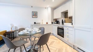 A modern kitchen with white cabinets, a dining table with chairs, and a coffee machine. - Modern Apartment By Historic Gloucester Quays (Gloucestershire)