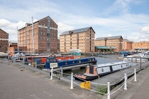 Zona junto a un canal con barcos amarrados, un edificio de almacén y edificios residenciales o comerciales aledaños.