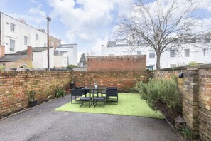 A patio area with a table and chairs, surrounded by a brick wall and a grassy patch.