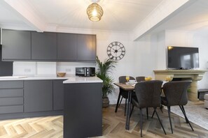 Eine moderne Küche mit grauen Schränken, einer zentralen Kochinsel und einem Essbereich mit Tisch und Stühlen. Eine Uhr und ein Flachbildfernseher sind an der Wand befestigt.