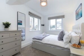 A bedroom with a bed, bedside table, dresser, windows, and a view of the outside.