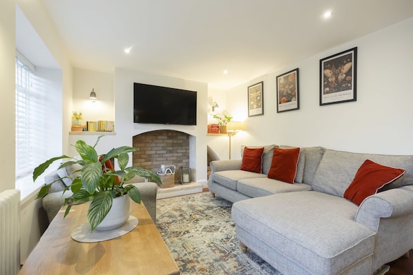 A living room with a fireplace, grey sofa, red cushions, a wooden coffee table, and a potted plant. - Charming Two Bedroom Apartment in Fairview (Gloucestershire)