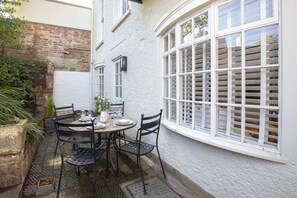 A patio with a table set for four, surrounded by chairs, against a white wall with a window featuring white shutters.