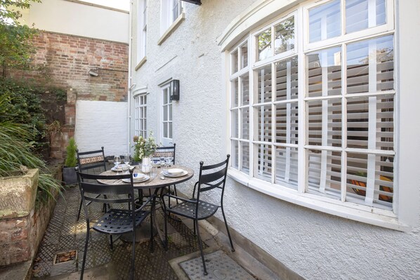 A patio with a table set for four, surrounded by chairs, against a white wall with a window featuring white shutters.