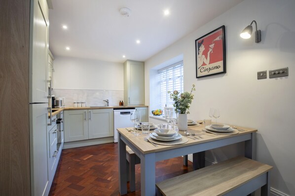 A modern kitchen with white cabinetry, a dining table set for four, and a window with blinds. - Charming Two Bedroom Apartment in Fairview (Gloucestershire)