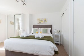 Un lit bien fait, avec du linge de lit blanc, un plaid vert et deux oreillers. Une petite table de chevet avec une lampe et un vase de fleurs se trouve à côté du lit. Un tableau est accroché au-dessus du lit, et une porte est visible sur la gauche.