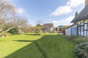 A well-maintained garden with a lawn and various plants, a traditional house with a thatched roof, and a clear blue sky. - Beautiful 4 Bedroom Cotswold Thatched Cottage (Twigworth)