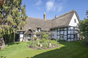 A traditional half-timbered house with a thatched roof, surrounded by a well-maintained garden and a large tree. - Beautiful 4 Bedroom Cotswold Thatched Cottage (Twigworth)