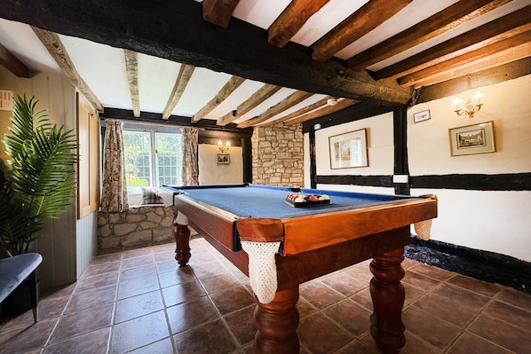 Une table de billard dans une pièce aux murs de pierre, aux poutres apparentes et dotée d’une fenêtre avec des rideaux.