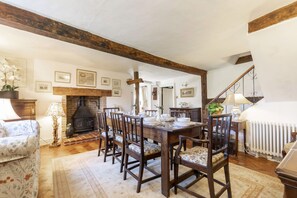 Une salle à manger avec une table en bois dressée pour six personnes, entourée de chaises. On aperçoit une cheminée au fond de la pièce, et des tableaux encadrés ornent les murs.
