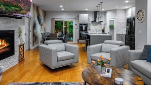 A modern living room with a fireplace, grey sofas, a wooden coffee table, and a kitchen in the background. - Game Room, Hot Tub, Cold Plunge, Sauna, Fire Pit (Asheville)