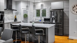A modern kitchen with stainless steel appliances, a marble island with bar stools, pendant lighting, and a large window with blinds. - Game Room, Hot Tub, Cold Plunge, Sauna, Fire Pit (Asheville)