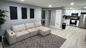 Een moderne woonkamer met een hoekbank, een getextureerd vloerkleed en een keuken met witte kasten en roestvrijstalen apparatuur.