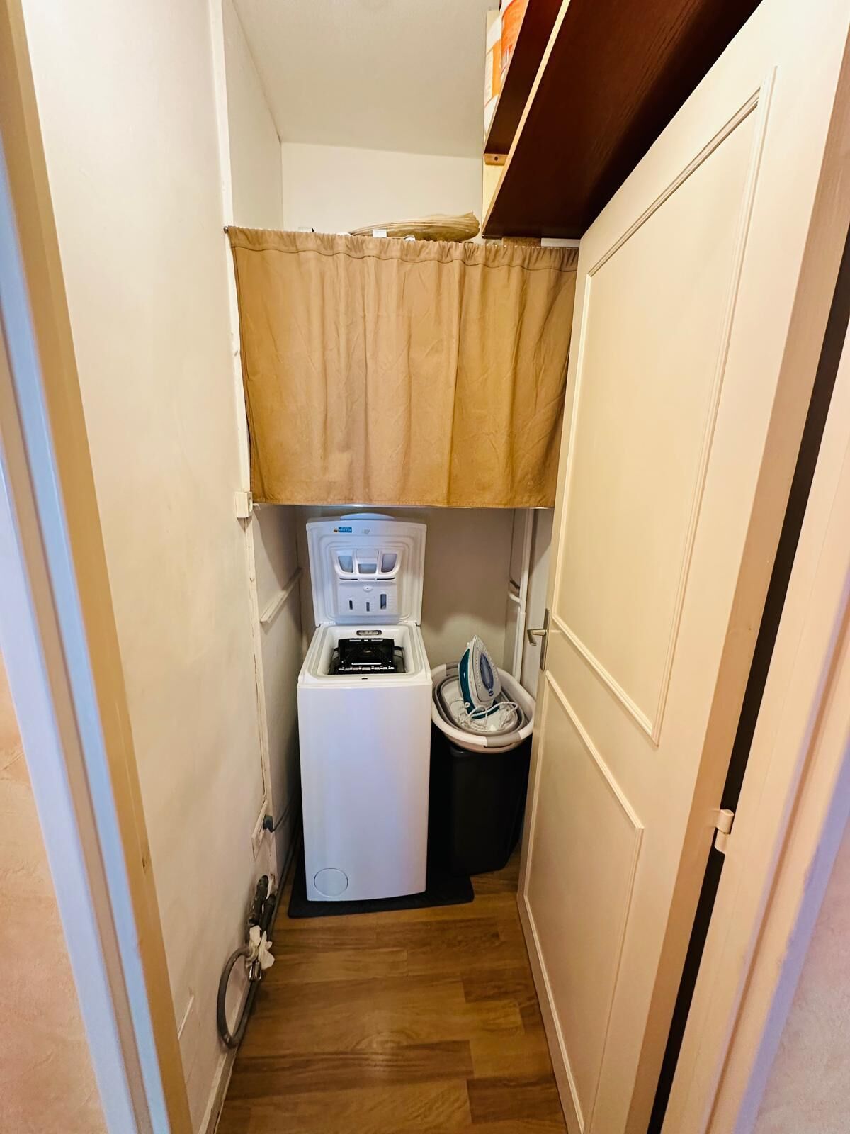 Een kleine wasruimte met een wasmachine en droger, een kleine prullenbak en een houten vloer.