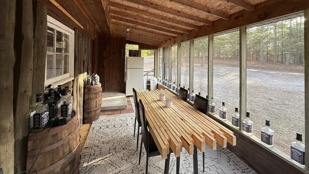 A rustic cabin interior with a long wooden table, bottles of Jack Daniel's whiskey, and a view of a forested area through large windows.