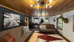 A cozy cabin interior with a wooden ceiling, a sofa, a bench, and a deer head mounted on the wall.