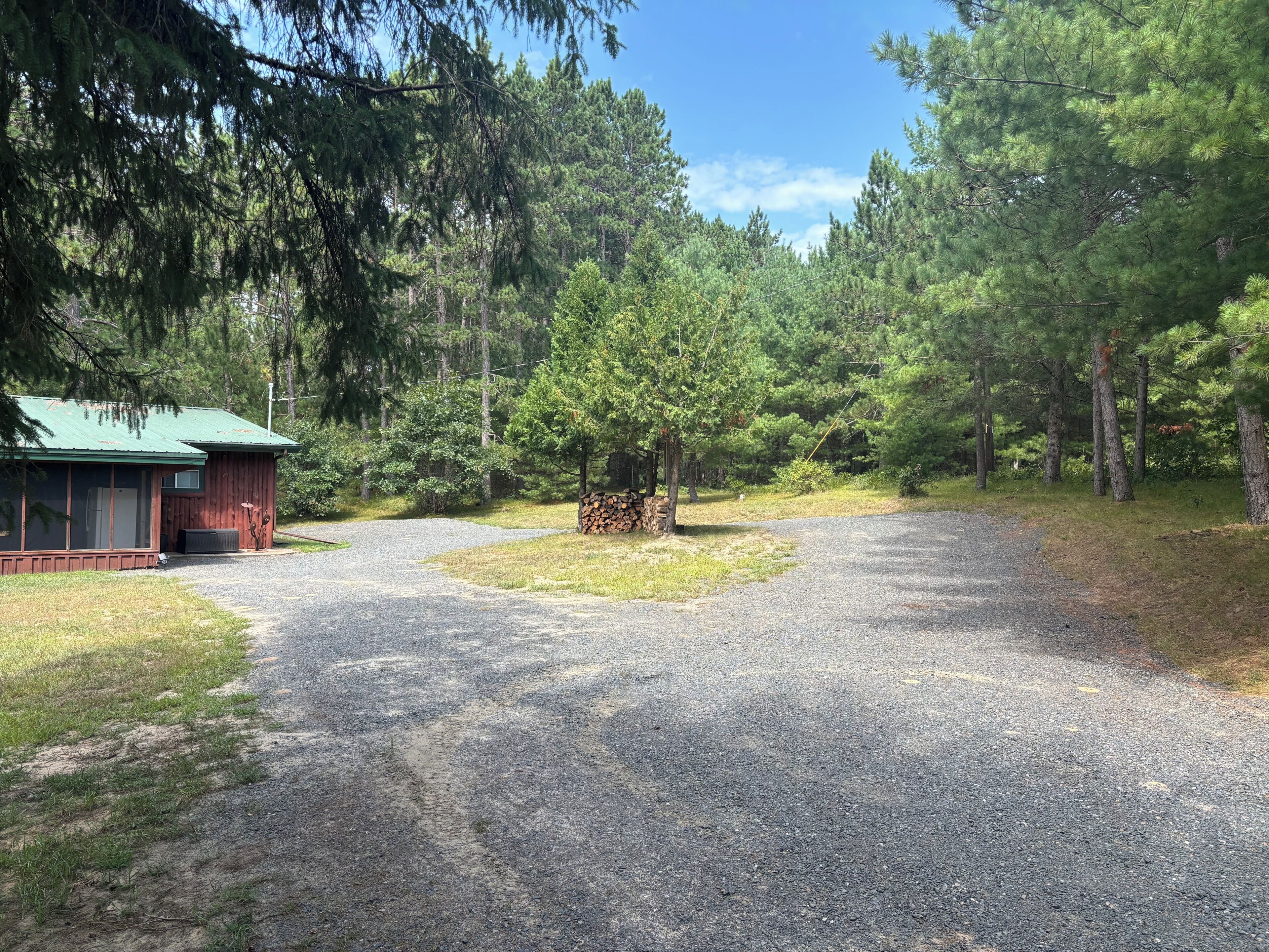 A gravel driveway leading to a cabin with a green roof, surrounded by dense forest.