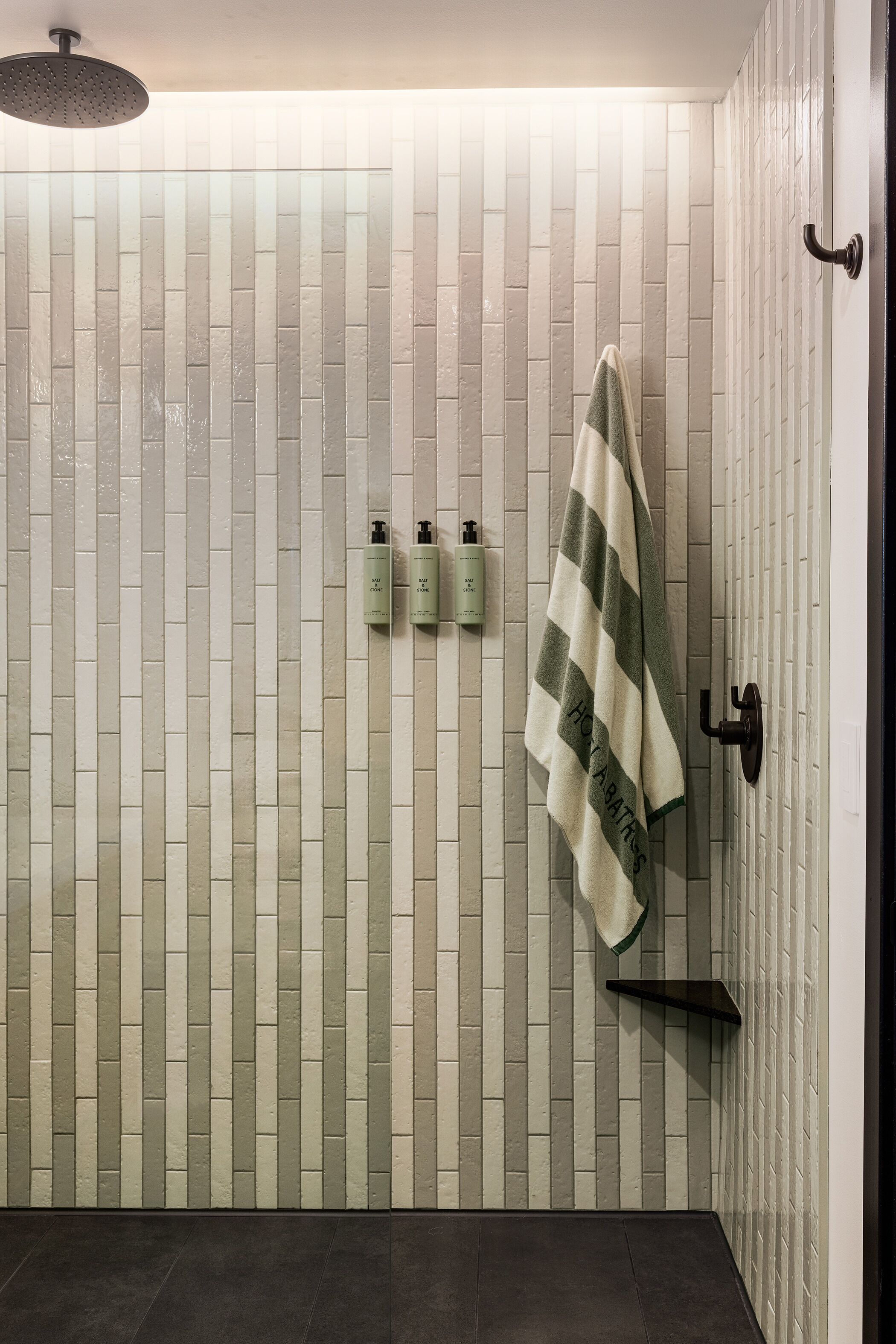 A bathroom with a tiled wall, a towel hanging on a rack, and three bottles of shampoo and conditioner on the wall.