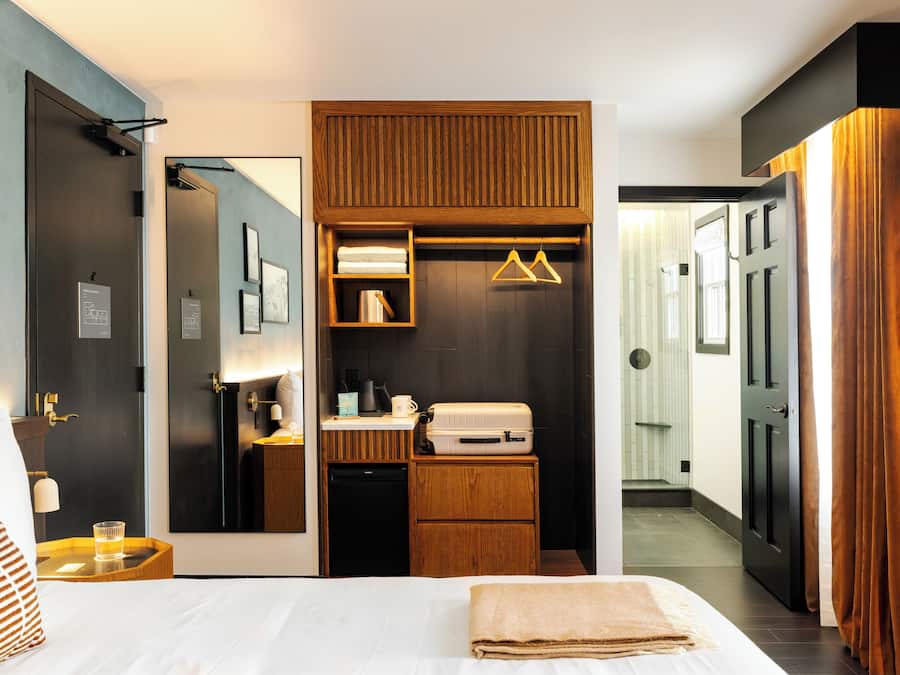 A hotel room with a bed, bedside table, mirror, wardrobe, and a small kitchenette area.