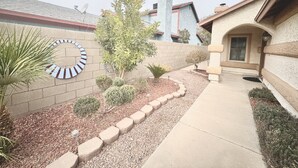 A residential area with a paved driveway, a landscaped garden with various shrubs and a palm tree, and a distinctive circular wall decoration. - Cozy 2-bedroom house in Henderson with WiFi, AC, Solar (Henderson)