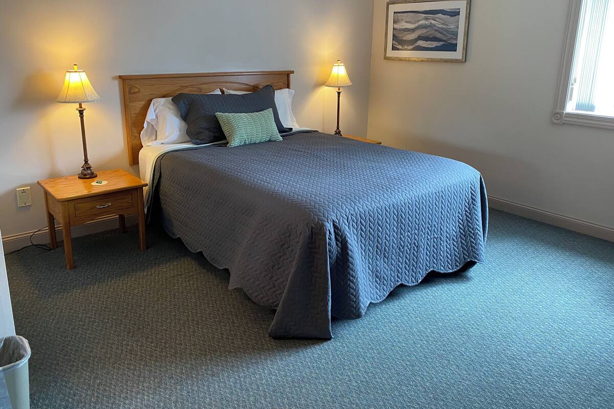A neatly made bed with a blue quilt, two bedside lamps, a small wooden nightstand, and a framed picture on the wall.