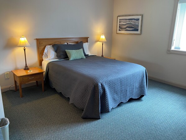 A neatly made bed with a blue quilt, two bedside lamps, a small wooden nightstand, and a framed picture on the wall. - Green Gables Inn (Lanesboro)