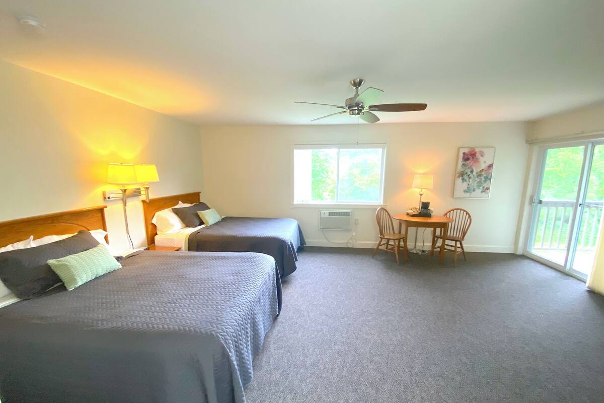 A hotel room with two beds, a ceiling fan, a small table with chairs, and a window with a view of greenery.