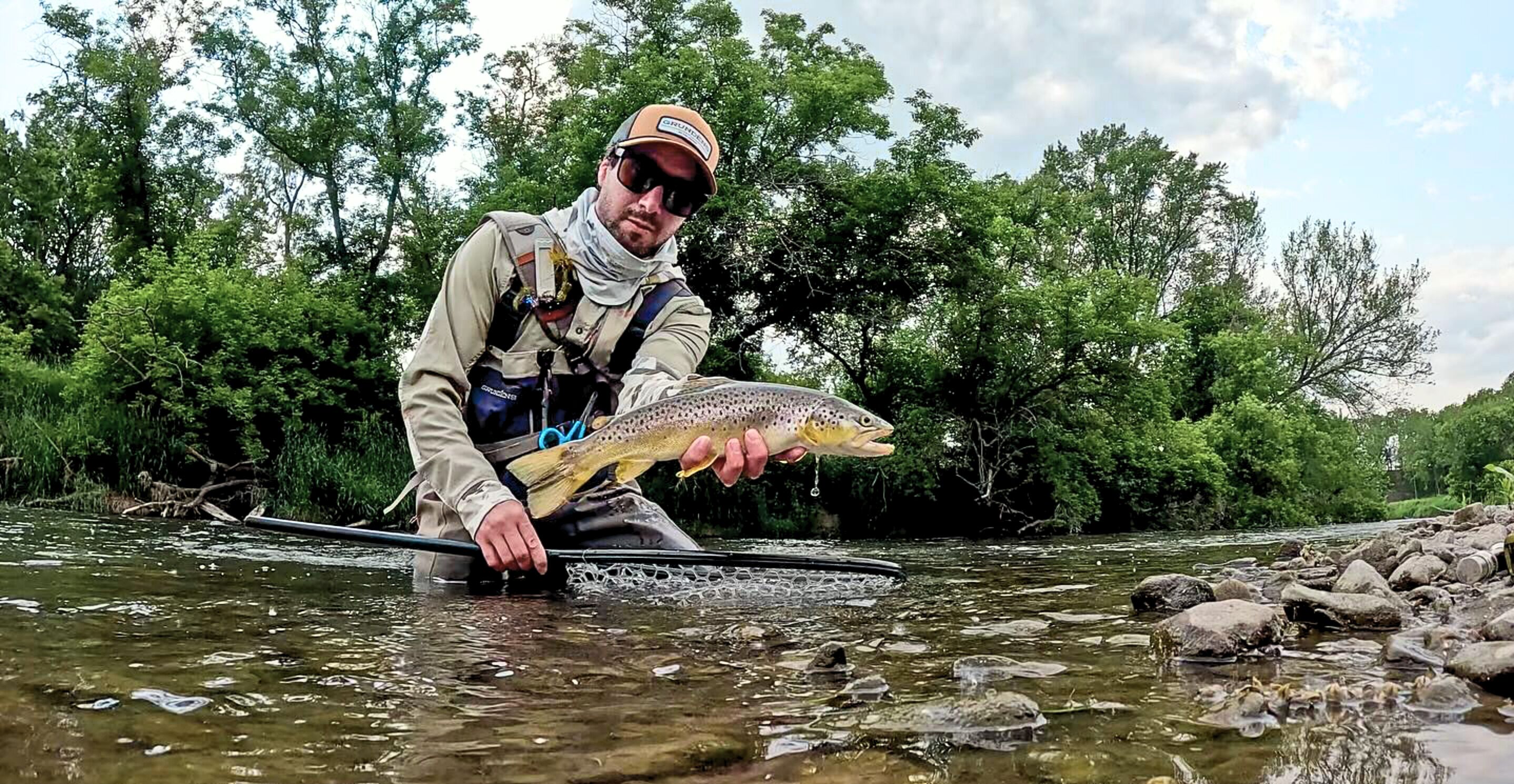 Une personne tenant un poisson dans une rivière, avec des rochers et des arbres en arrière-plan.