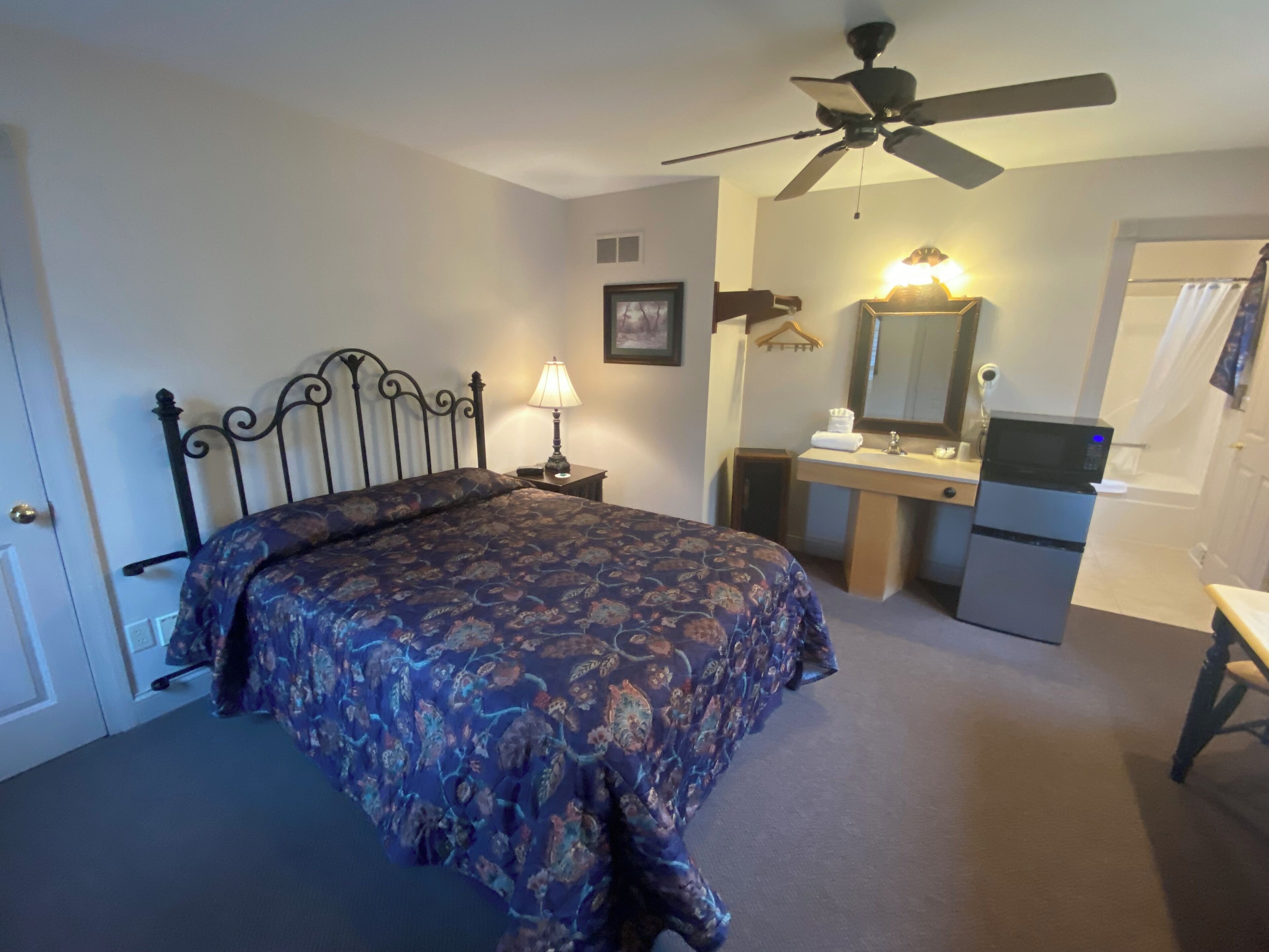 A bedroom with a bed, a nightstand, a desk, a mirror, a ceiling fan, and a bathroom visible through an open door.