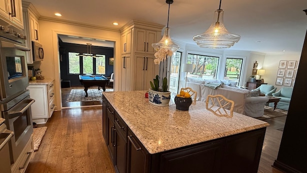 A spacious kitchen with granite countertops, dark cabinetry, and a central island. There is a pool table in the background and a dining area with a light blue sofa.
