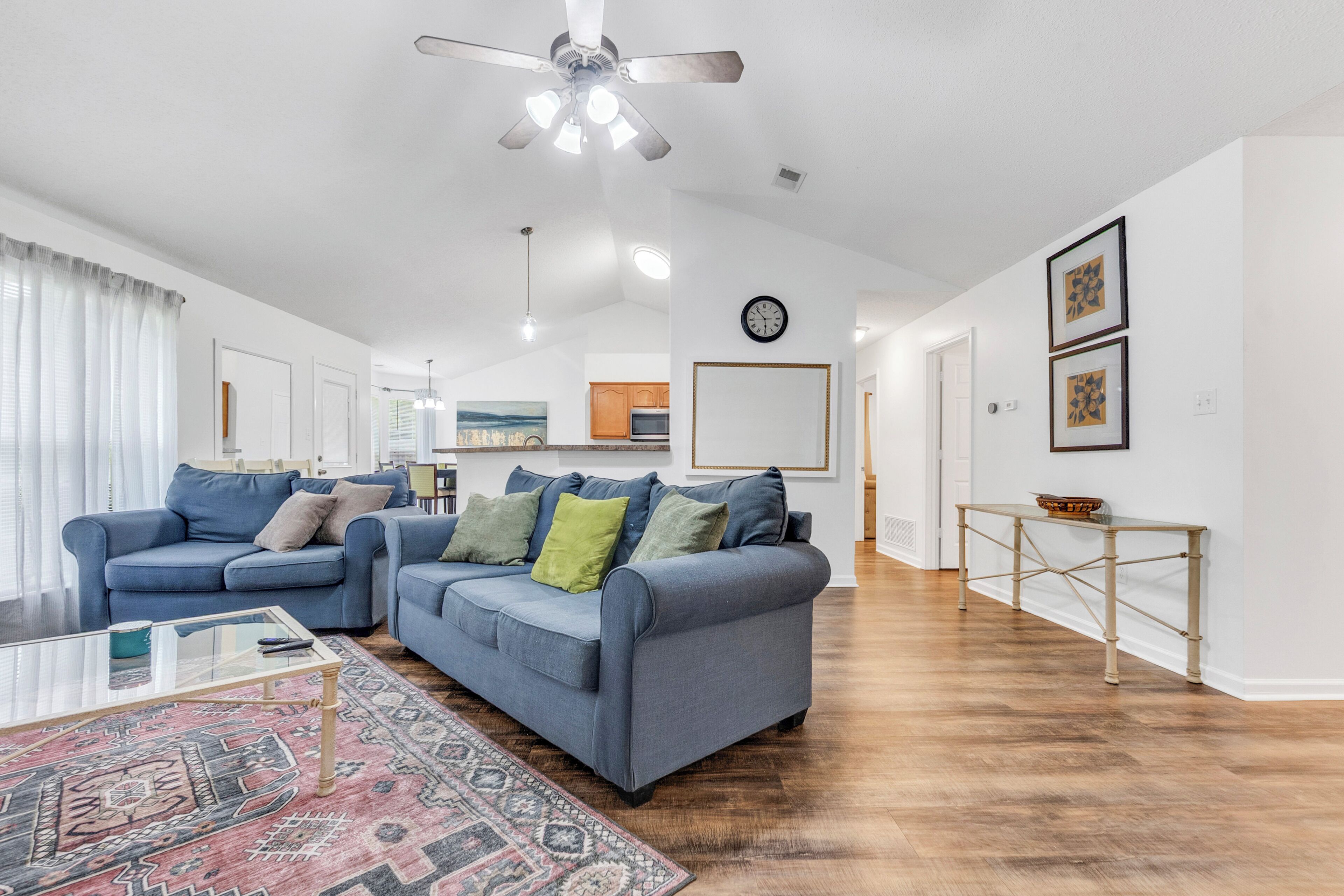 A living room with two blue sofas, a glass coffee table, a patterned rug, a ceiling fan, and a kitchen in the background.