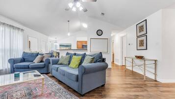 A living room with two blue sofas, a glass coffee table, a patterned rug, a ceiling fan, and a kitchen in the background.