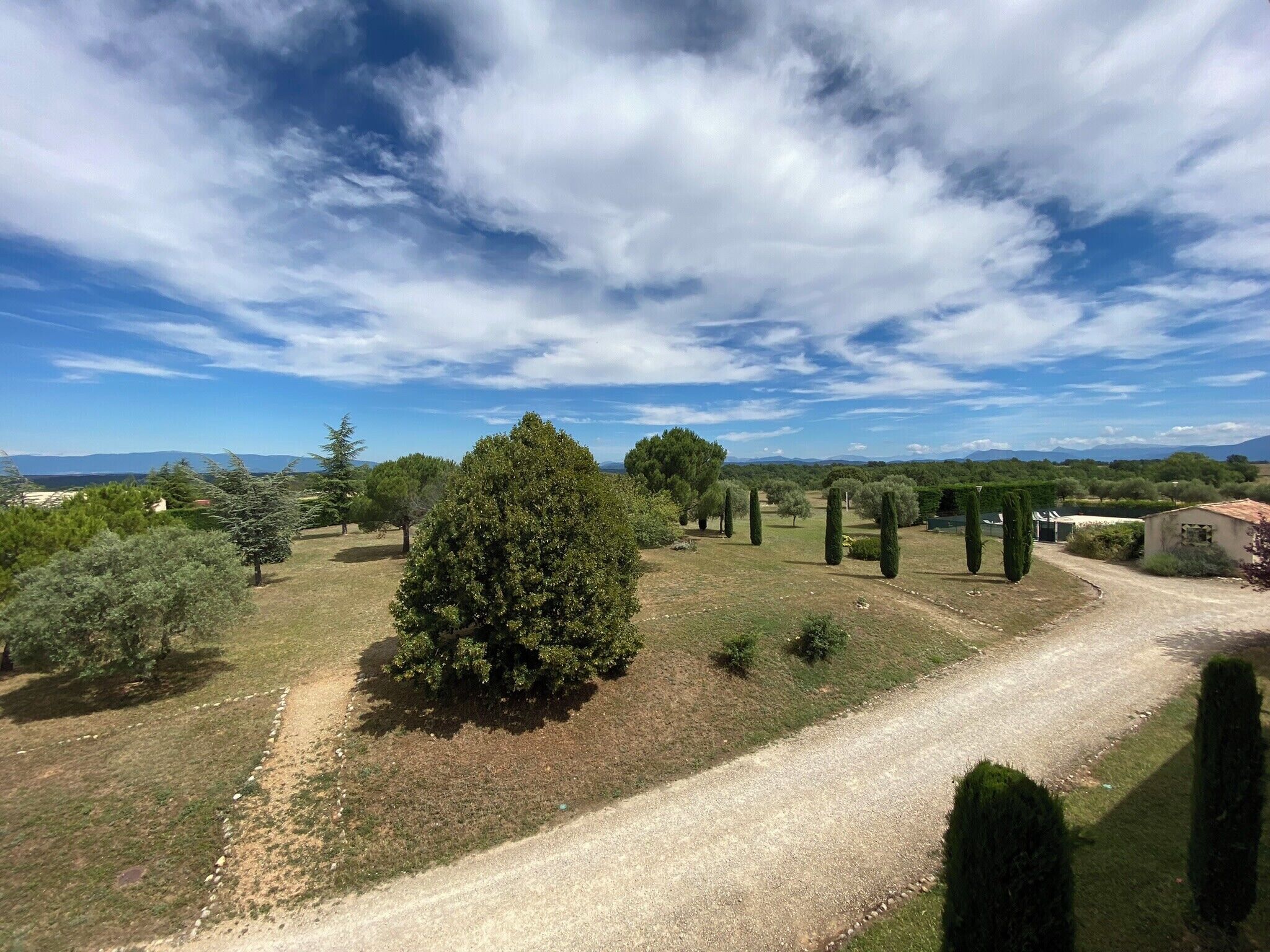 Paisaje rural con un sendero de grava, varios árboles y montañas lejanas bajo un cielo parcialmente nublado.