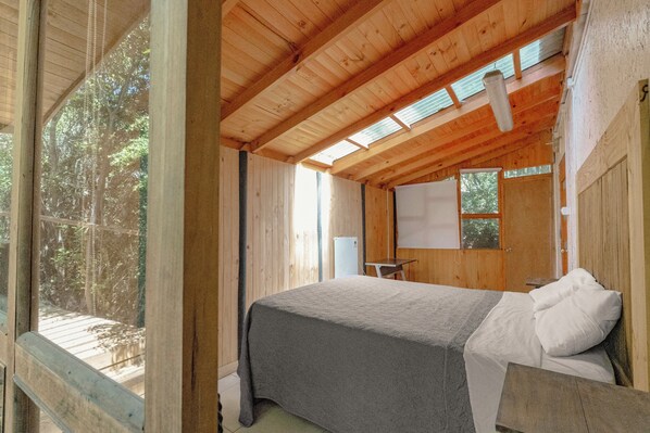 A wooden cabin bedroom with a bed, a desk, and a window overlooking a forest. - ecorefugio spa (Colbun)