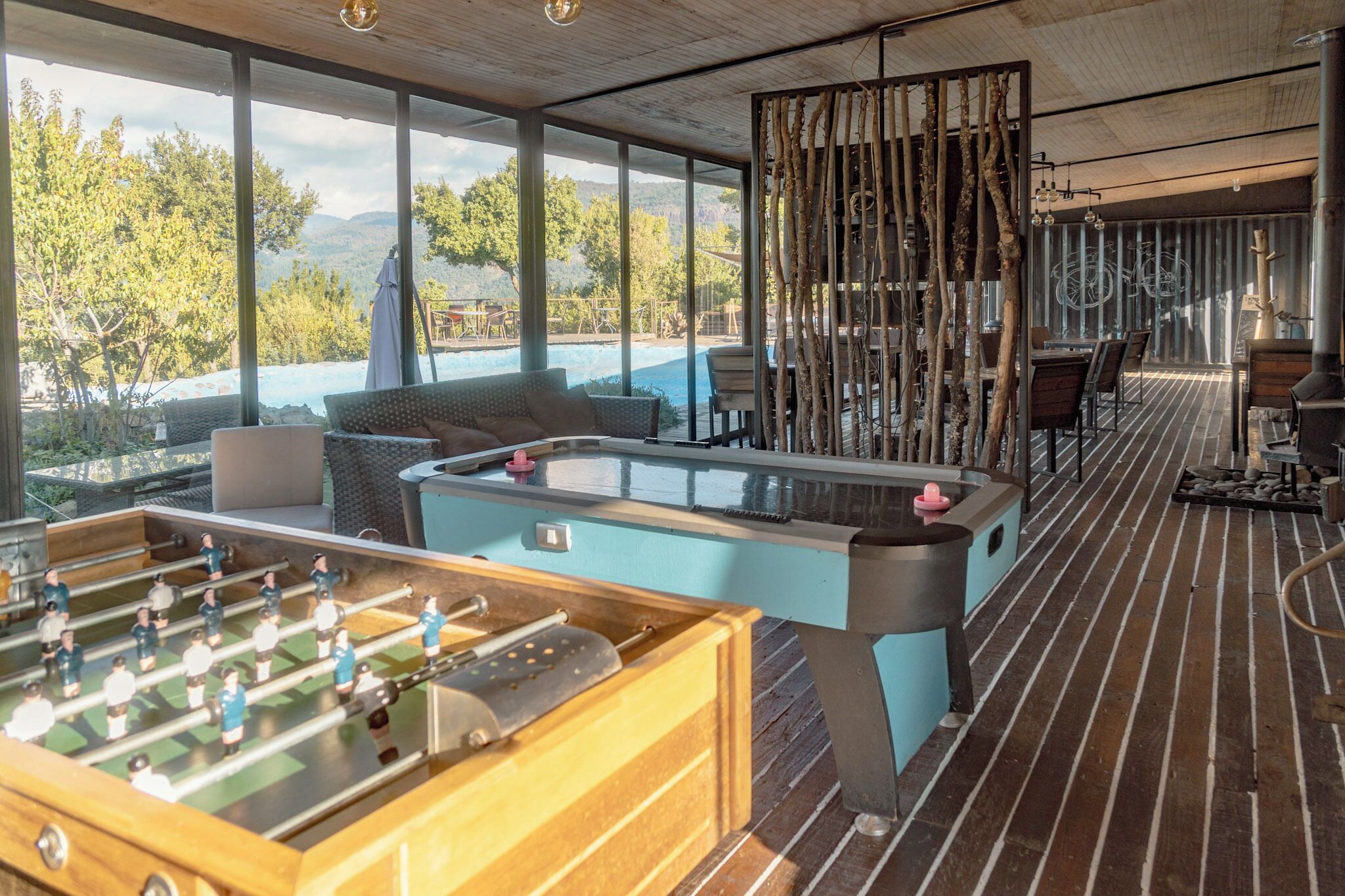 A pool table and foosball table in a spacious room with large windows overlooking a pool and mountains.