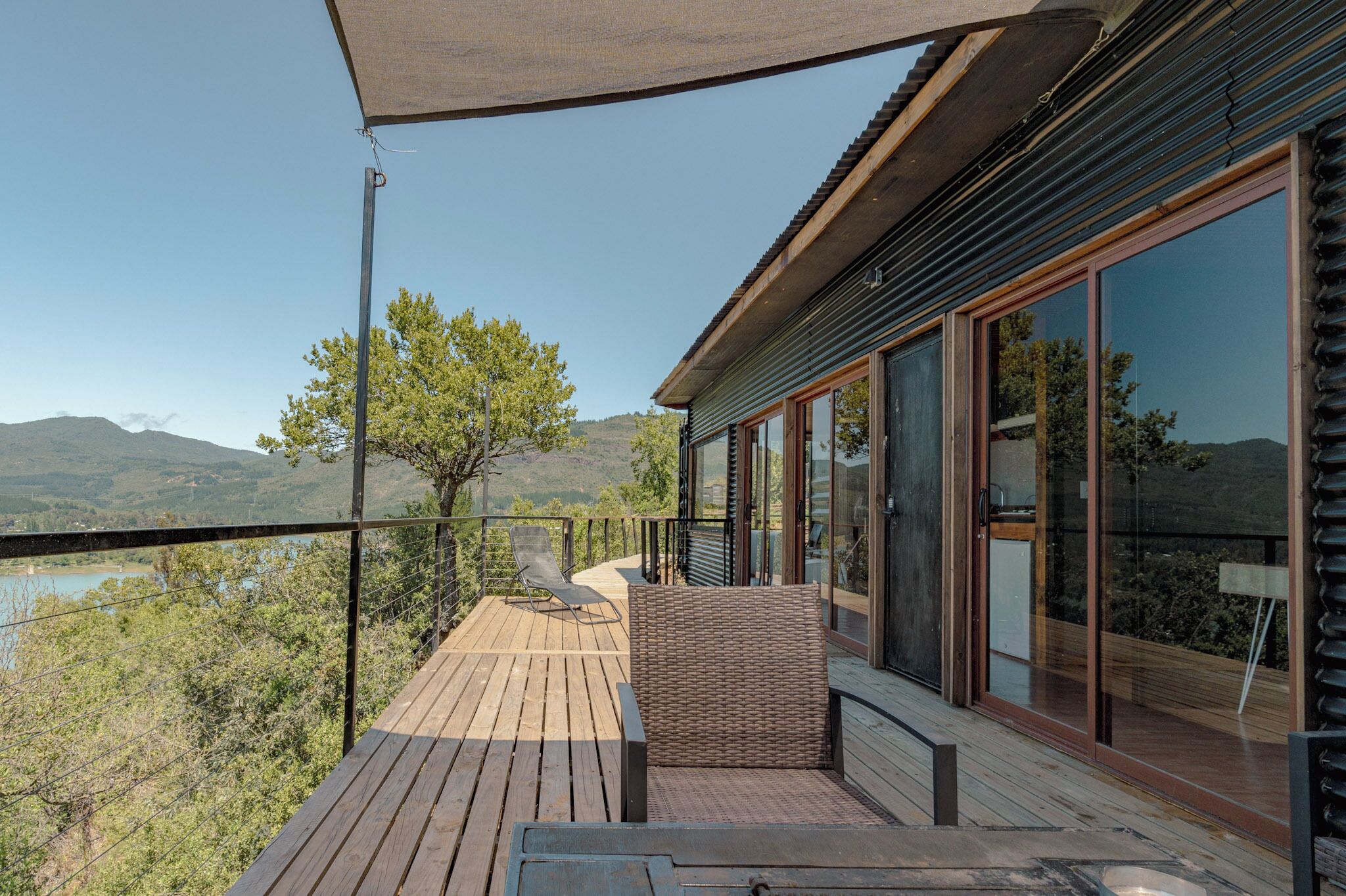 A wooden deck with a wicker chair and a table, overlooking a lake and hills.