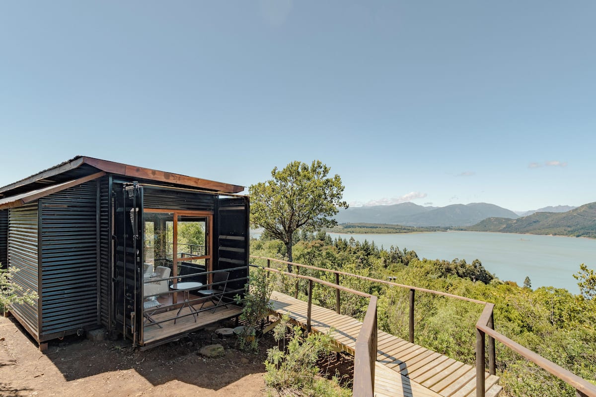 A cabin with a glass door, a deck, and a view of a lake and mountains.