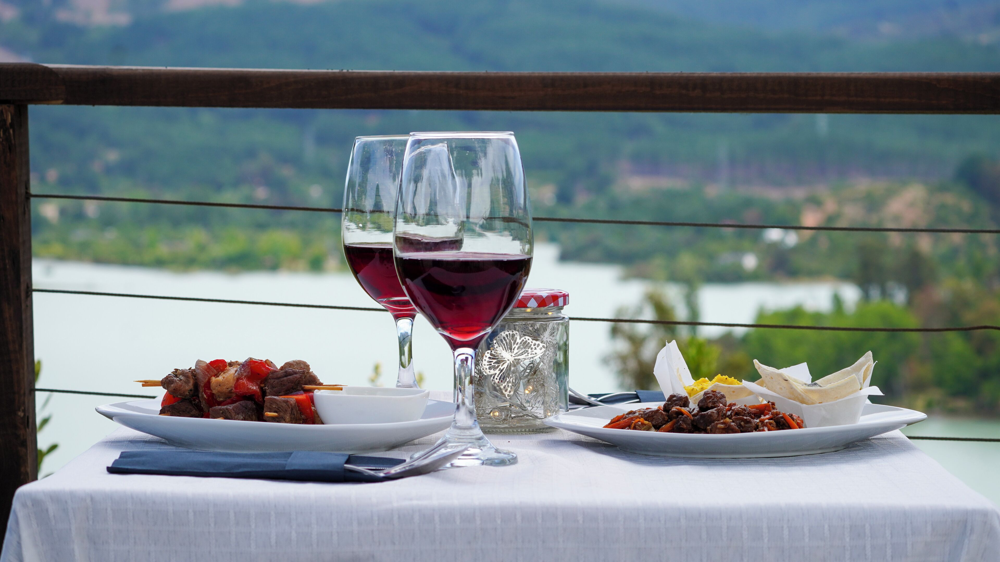 Una mesa con dos copas de vino tinto, brochetas de carne y un tarro de especias, frente a una vista panorámica de un lago.