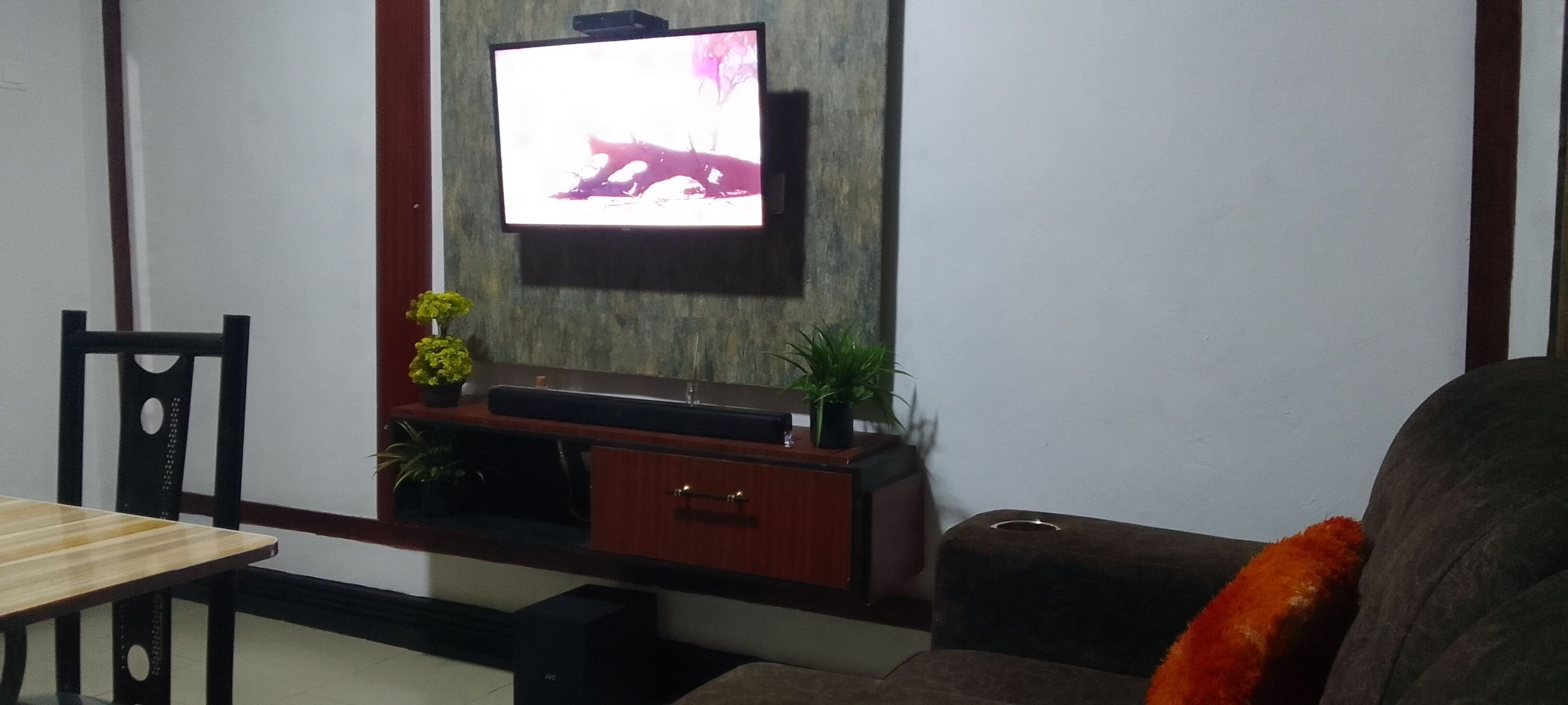 A living room with a flat-screen TV mounted on the wall, a wooden coffee table, a brown sofa, and a small plant on the side table.