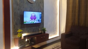 A living room with a flat-screen TV mounted on the wall, a dark brown sofa, a wooden entertainment unit with a plant, and a window with curtains. - MVH Apartment in Sunyani with free WiFi, AC and fully furnished kitchen. (Sunyani)