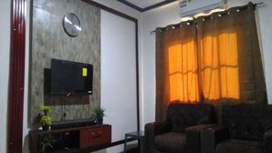 A living room with a flat-screen TV mounted on a wall, a clock, a sofa, and a window with curtains. - MVH Apartment in Sunyani with free WiFi, AC and fully furnished kitchen. (Sunyani)