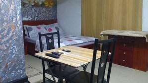 A small room with a bed, a desk with a chair, a table with a tablecloth, and a wall with a floral pattern. - MVH Apartment in Sunyani with free WiFi, AC and fully furnished kitchen. (Sunyani)