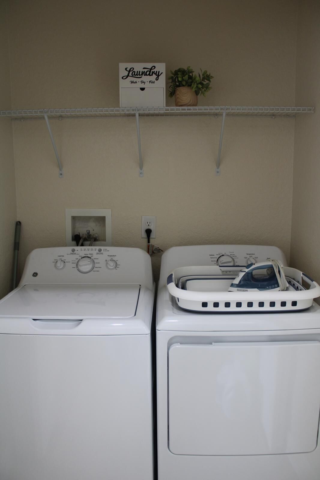 A laundry room with two front-loading washing machines and a shelf above holding a sign and a potted plant.