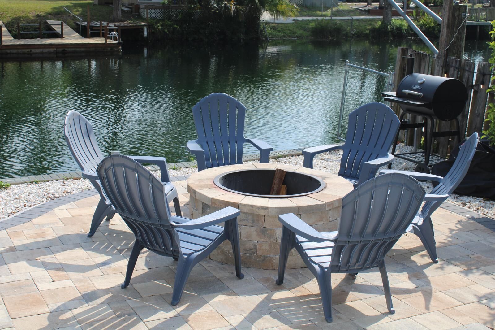 Un hogar circular de piedra rodeado por cuatro sillas Adirondack en un patio empedrado junto a un cuerpo de agua.