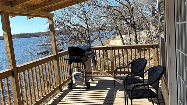 A wooden deck with a grill, chairs, and a view of a lake.