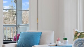 Une pièce avec un fauteuil blanc et un canapé bleu, tous deux dotés de coussins. On y trouve une petite table sur laquelle sont posés une plante et un objet décoratif. Une fenêtre offre une vue sur les arbres.