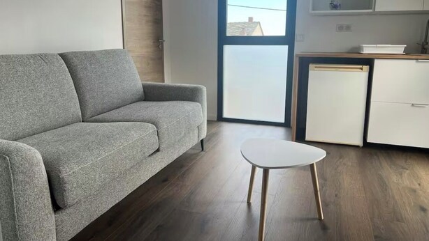 Una sala moderna con un sofá gris, una mesita blanca y una cocina con armarios blancos y un ventanal.